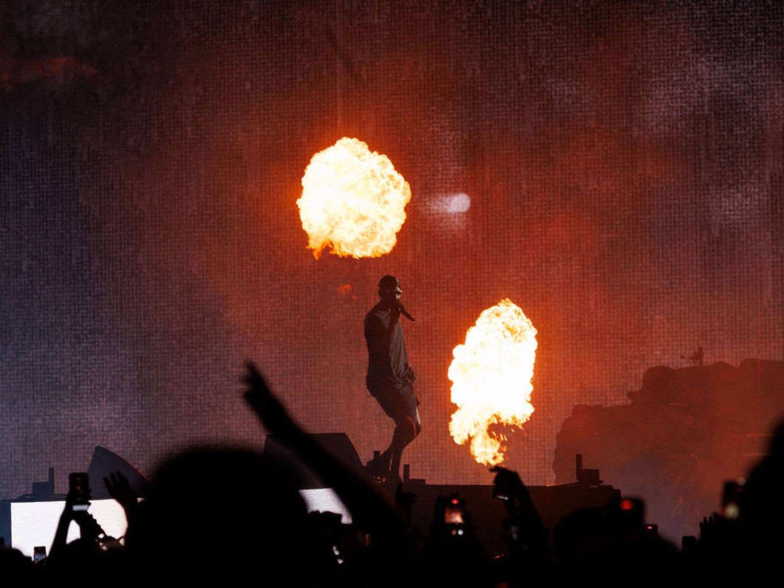 2024 - Travis Scott Travis Scott performing on stage at Suncorp Stadium in 2024 with pyrotechnics going off behind him. Fans arms can be seen in the foreground.